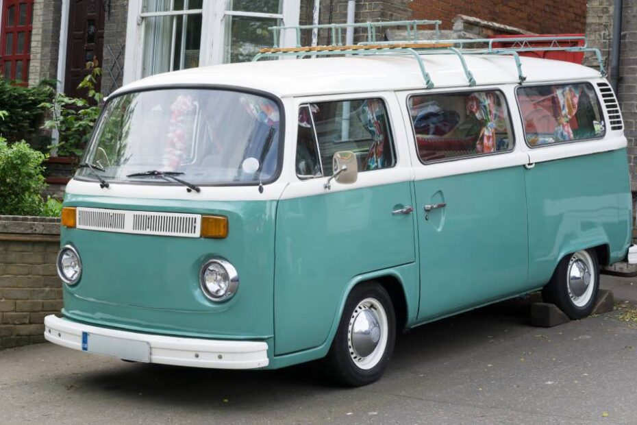 A light blue classic Volkswagen camper van with white trim sitting on the driveway of a brick townhouse.