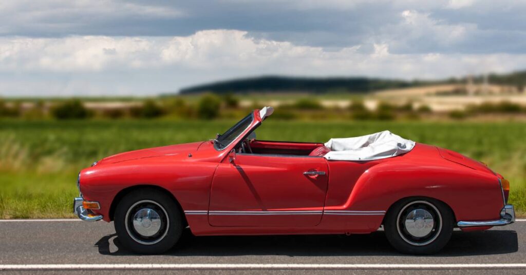 A red Karmann Ghia Cabrio sits on a road surrounded by a beautiful countryside landscape with dark clouds above.