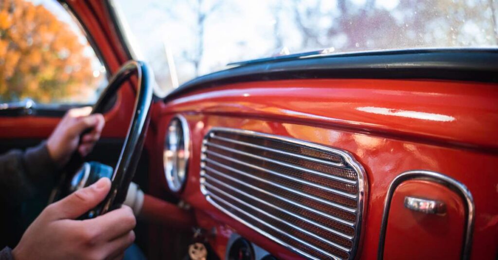 A driver has both hands on a black steering wheel. The vintage car's entire interior is red with chrome trim.