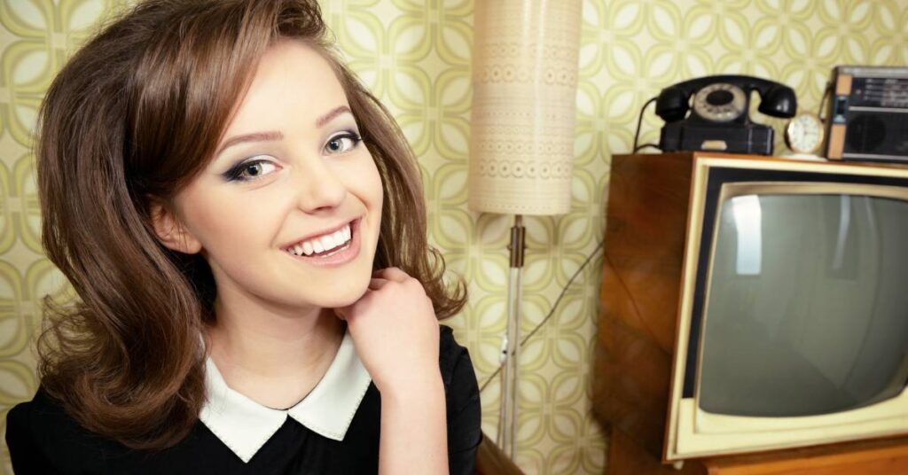 A young woman with wavy brown hair wears a black dress with a white Peter Pan collar. She’s smiling directly at the camera.