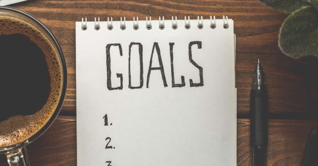 A white notepad sits on a wooden table. The word "Goals" appears at the top, and the numbers one, two, and three appear below.