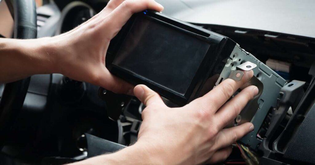 A pair of hands hold a car radio sound system in the front of a car. They're installing it on the dashboard.