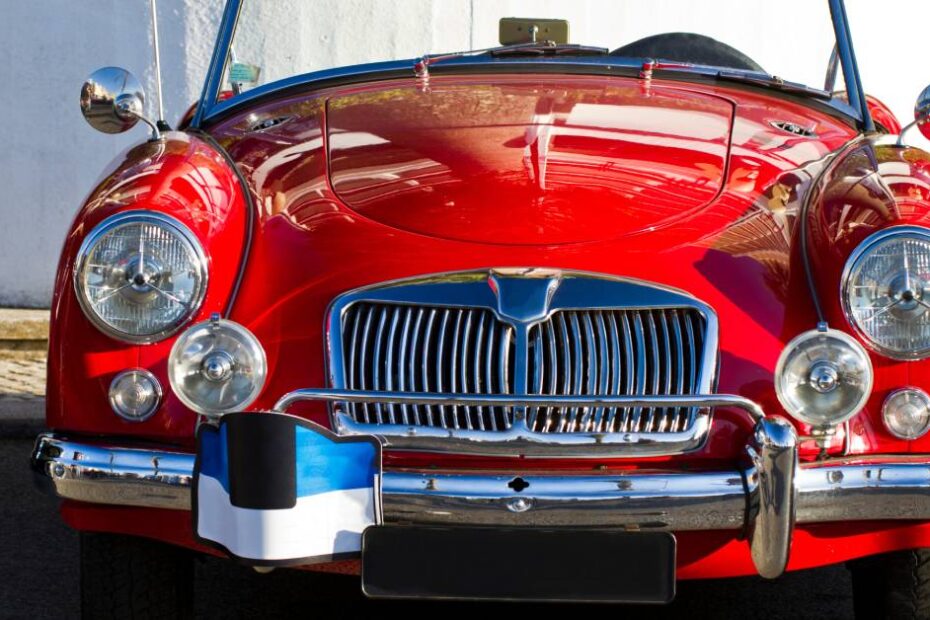 A close-up of a red vintage convertible with chrome detailing and vintage lights and mirrors. The car sits on a paved road.