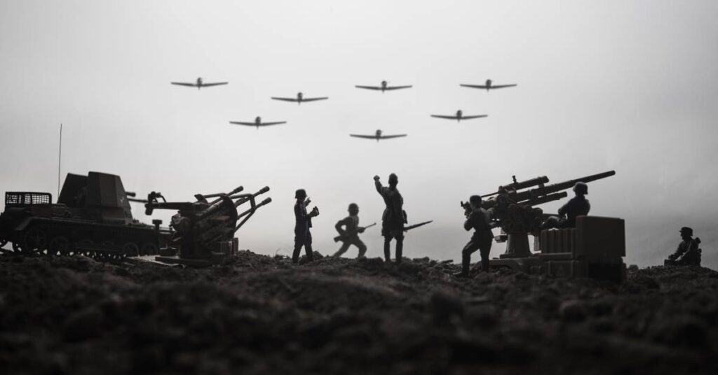 Soldiers standing on a hill with tanks and cannons. They are looking up as seven planes fly above them.