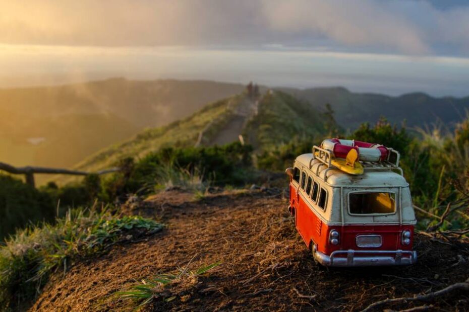 A miniaturized orange and white Volkswagen Bus sits on top of a mountain. The cloudy skies and other peaks are in the distance.