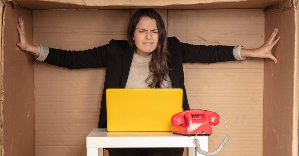 A woman sits at a table and stretches her arms inside a cardboard box. On the table are a red phone and a yellow laptop.