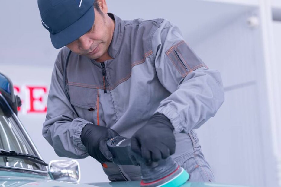 A man wearing a grey mechanic’s uniform and blue hat uses a polishing tool on a light blue classic vehicle.