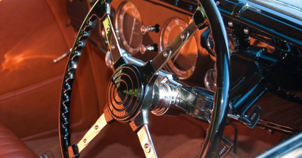 A classic car has a black steering wheel, black dashboard, and orange upholstery. The interior looks shiny and polished.
