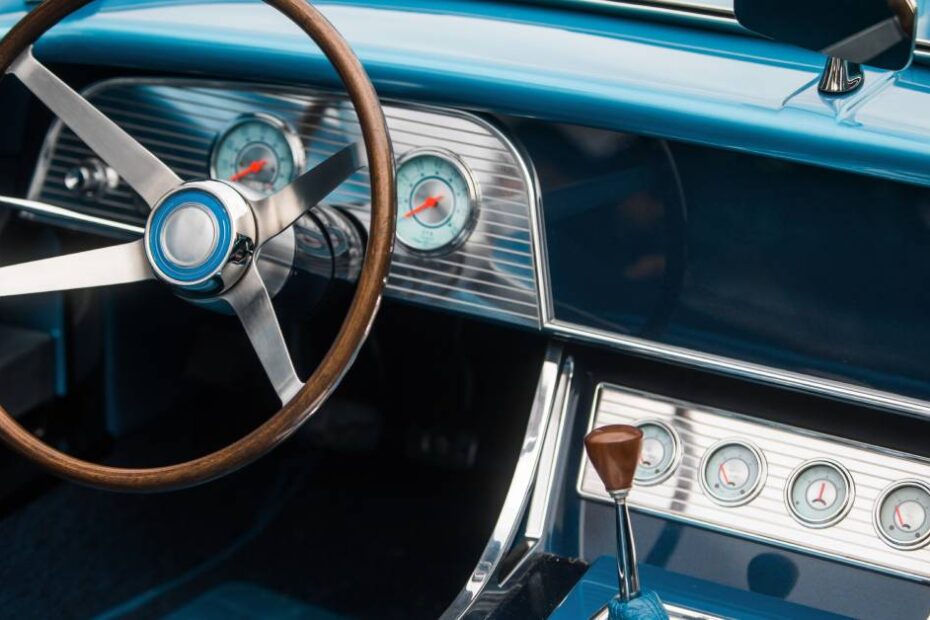 The inside of a classic car’s dashboard has a blue interior, a wooden steering wheel, and a silver dashboard.