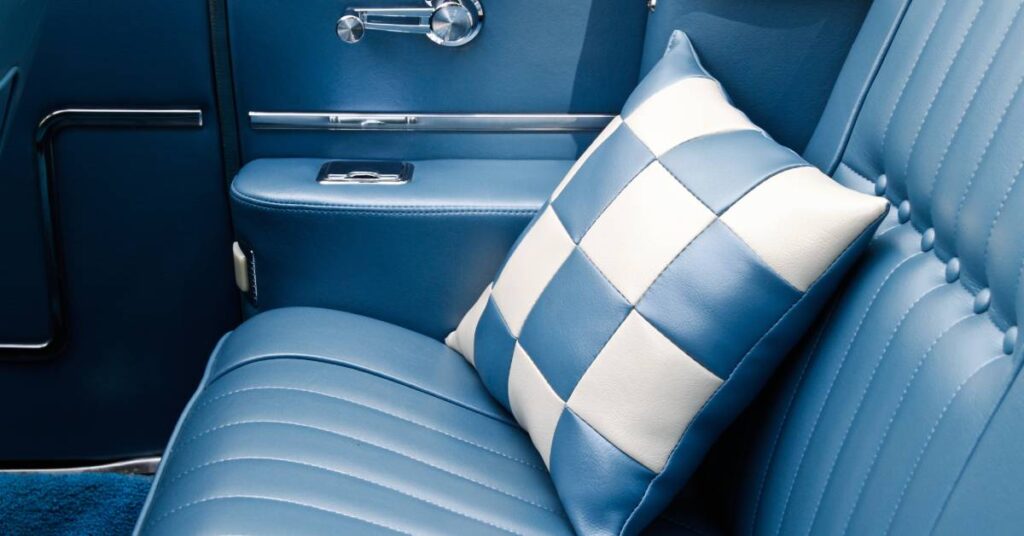 A classic car's interior has blue leather seats and upholstery. The backseat has a blue and white checkered pillow.
