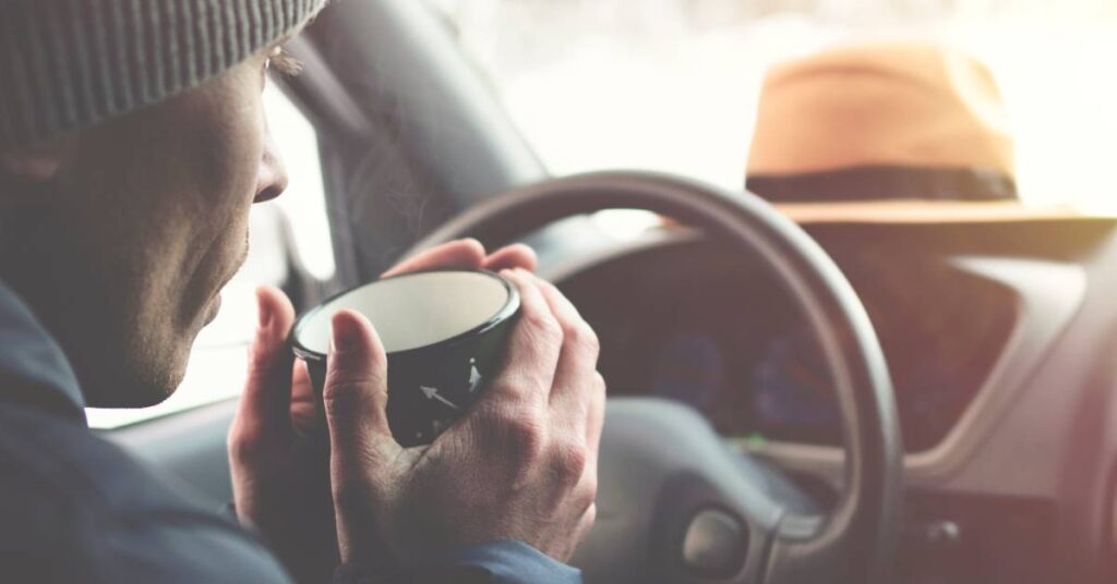 A man wearing a grey beanie holds a cup with both hands while behind the wheel of a car. Snow is outside the car’s window.