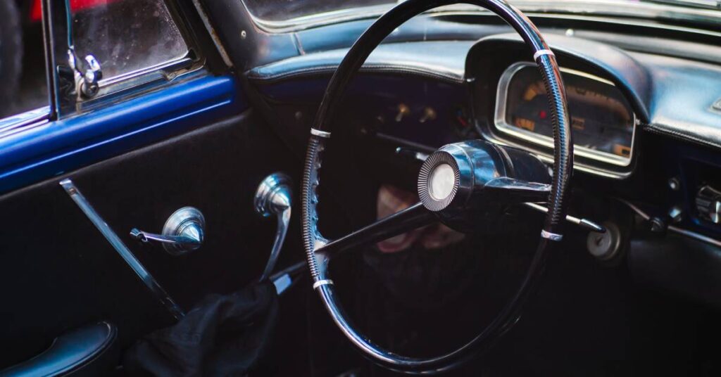 A close-up view of the black leather interior of a vintage automobile. The vintage steering wheel and dashboard are visible.