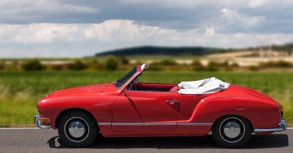 A red Karmann Ghia Cabrio is parked on a road with its top down. A green field and cloudy skies are visible behind the car.
