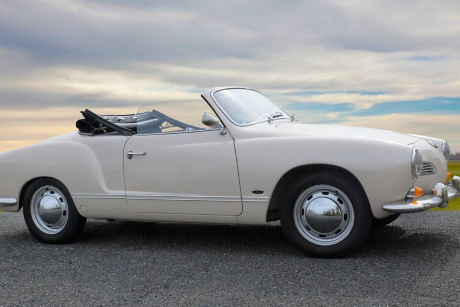 A white Karmann Ghia Oldtimer sits on a gravel road with its top down. There is a green field in the background.