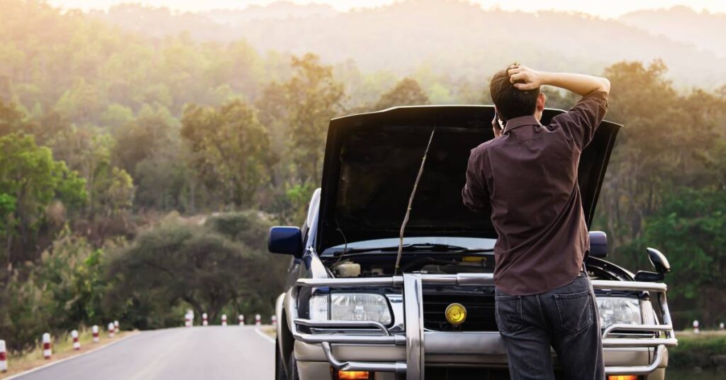 A man talks on a cell phone while standing on the side of the road. He stands in front of a car with the engine propped open.