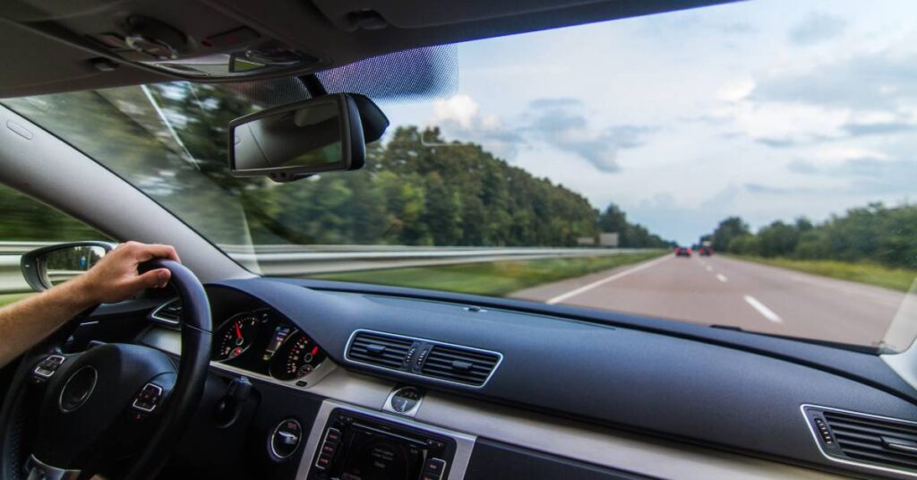 A man’s left-hand holds the steering wheel as he drives on a highway. The car has a black, modern interior.