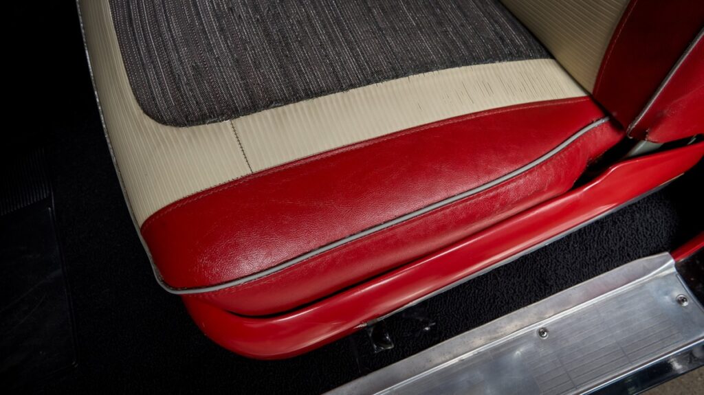 A vehicle seat with red and tan leather has a grey fabric center. A thin piece of metal is beside the seat.