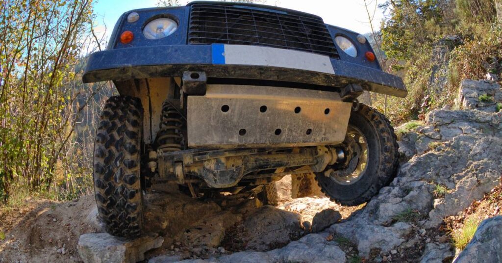 A close-up shot of a vehicle attempting to drive over a rocky terrain. Trees surround the car on each side.