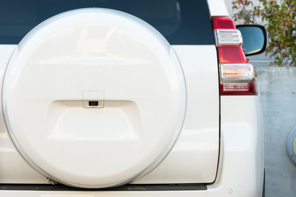 A white metal spare wheel cover is on the back of a white SUV. The car’s headlights and right rear-view mirror are visible.