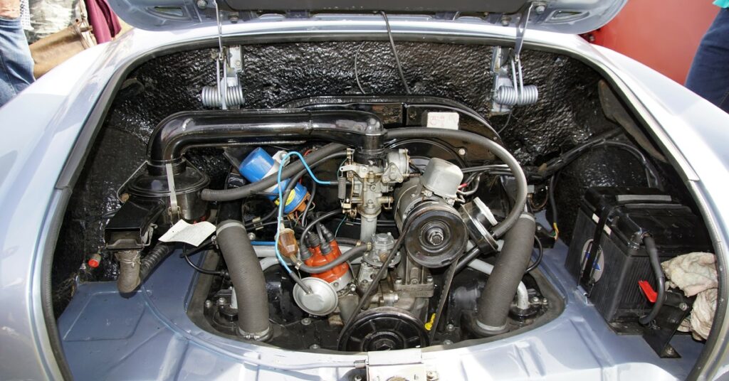 The engine of a silver Volkswagen Karmann Ghia revealing cables, hoses, belts, and the engine block.