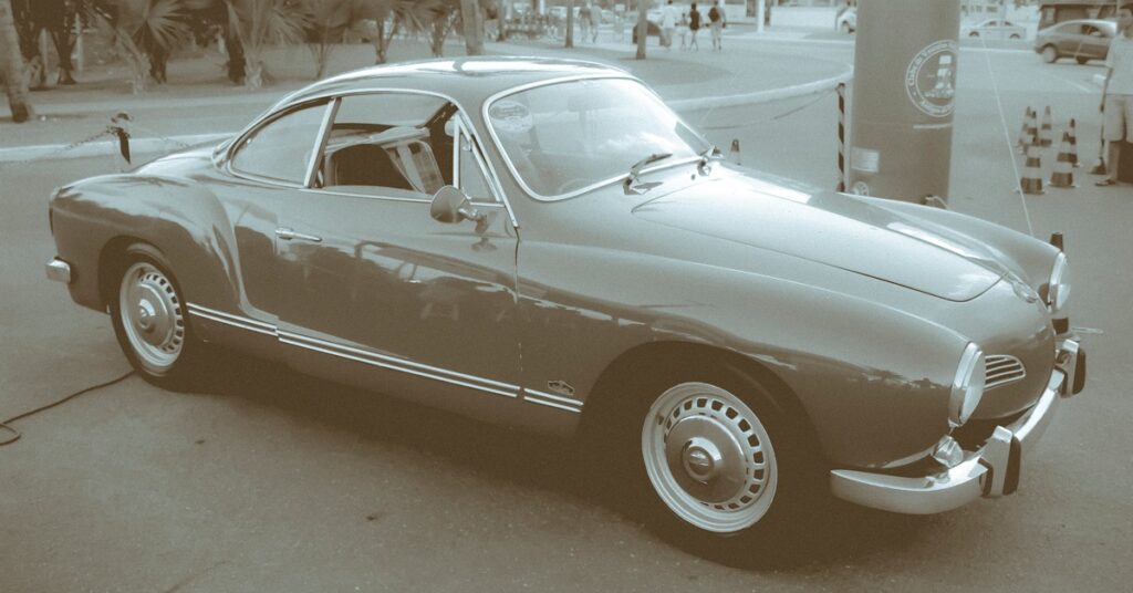 A Volkswagen Karmann Ghia filtered in a black-and-white photo setting with people and trees seen in the background.