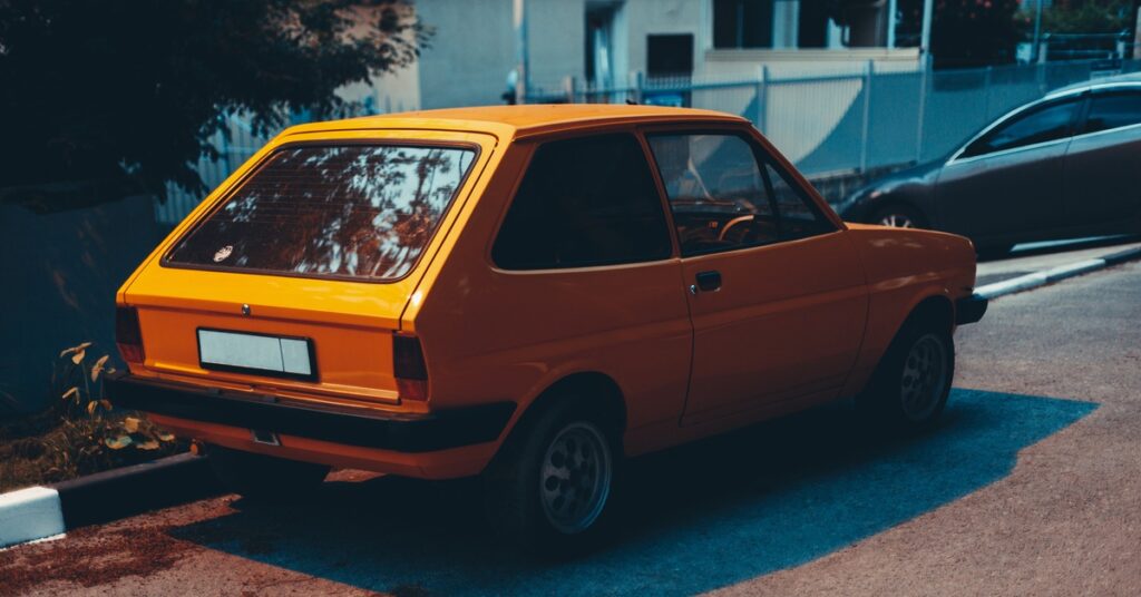 A yellow car with a sloped back parked on the side of a residential street at night. Another car is in a nearby driveway.