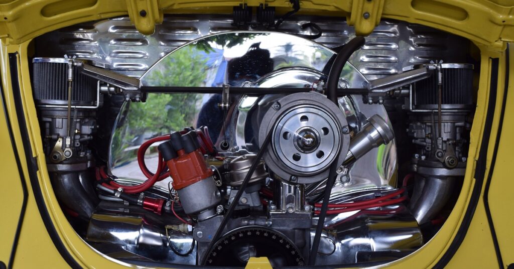 The exposed engine of a dark-yellow Volkswagen Beetle with a reflective metal surface and numerous belts and cables.