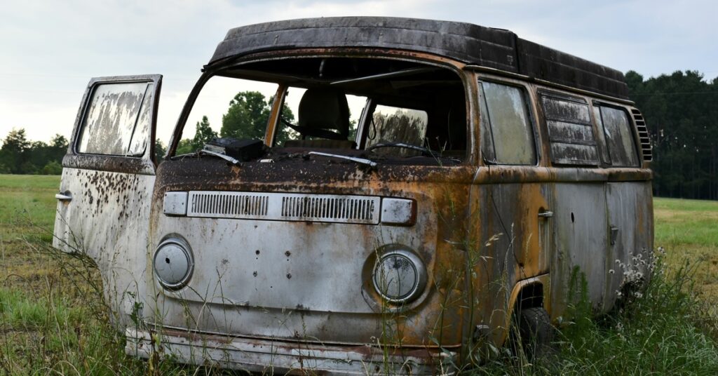 A rusted Volkswagen Bus abandoned in the middle of a field with the passenger door open and tall grass surrounding it.