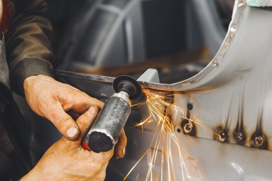 A using a handheld grind to smooth out the rusted edges of a silver car's chassis a sparks fly in multiple directions.