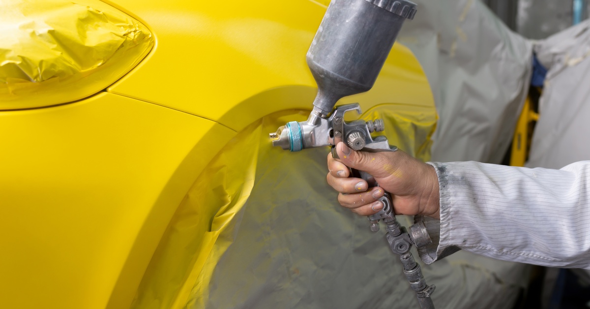 A person wearing white coveralls using a spray gun to paint the fenders of a car with yellow paint.