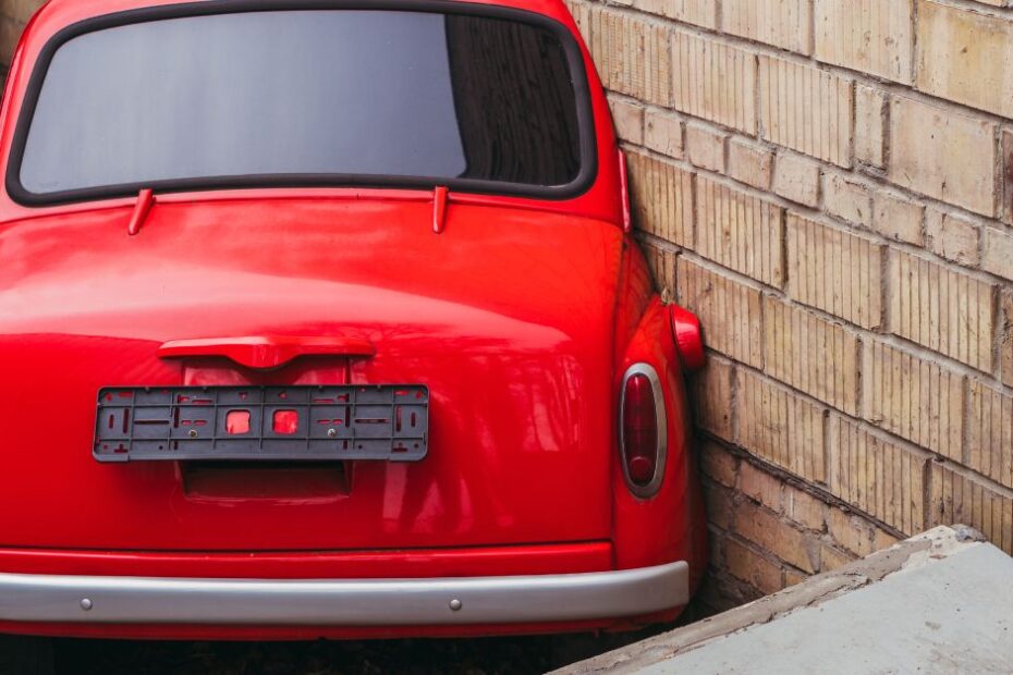A red Volkswagen Beetle parked diagonally against a brown brick wall with numerous dead leaves scattered around it.