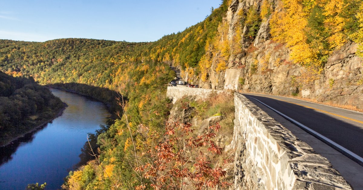 A cliffside road on the Catskills Scenic Byway with the Delaware River and green and yellow trees parallel to its path..