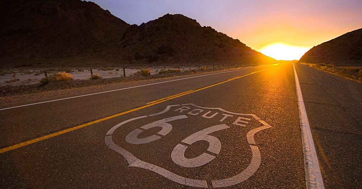 A long road with "Route 66" in a shield painted on its surface. The sun is setting between two small mountains on the horizon.
