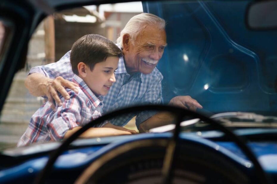 An older adult man and a young boy looking under a vintage car hood and examining the engine on a sunny day.