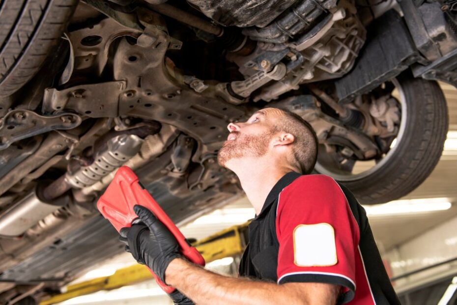 A bearded male mechanic viewing the underside of a raised vehicle and taking notes in a small red tablet.