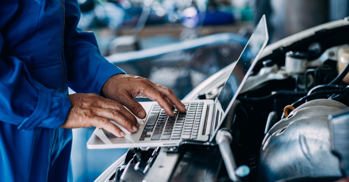 A mechanic in a blue jumpsuit stands inside a repair shop using a laptop perched on a car's engine bay.