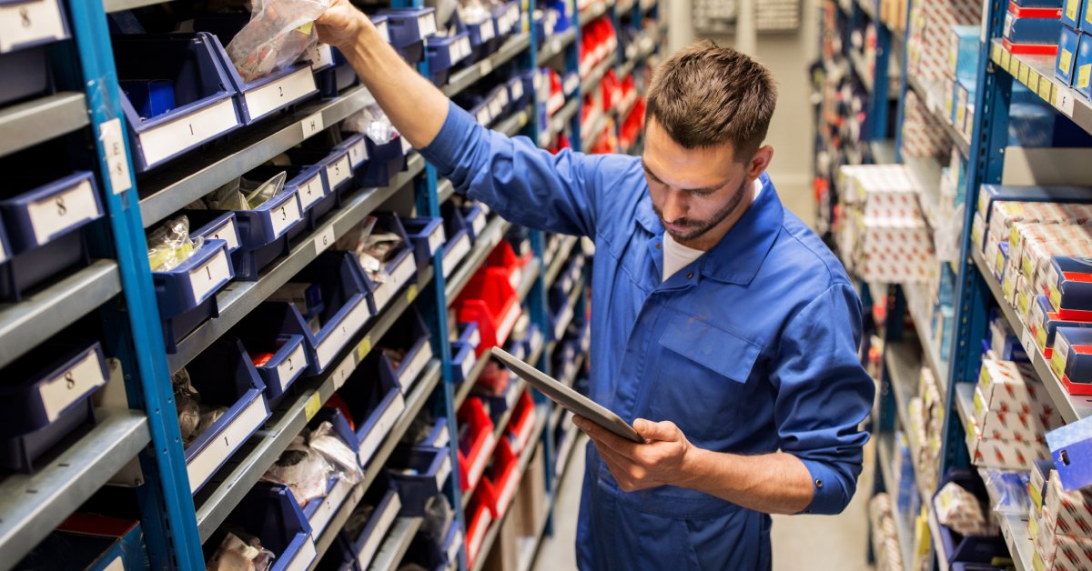 A mechanic in a jumpsuit grabbing auto parts from a high, crowded shelf while consulting a silver tablet.