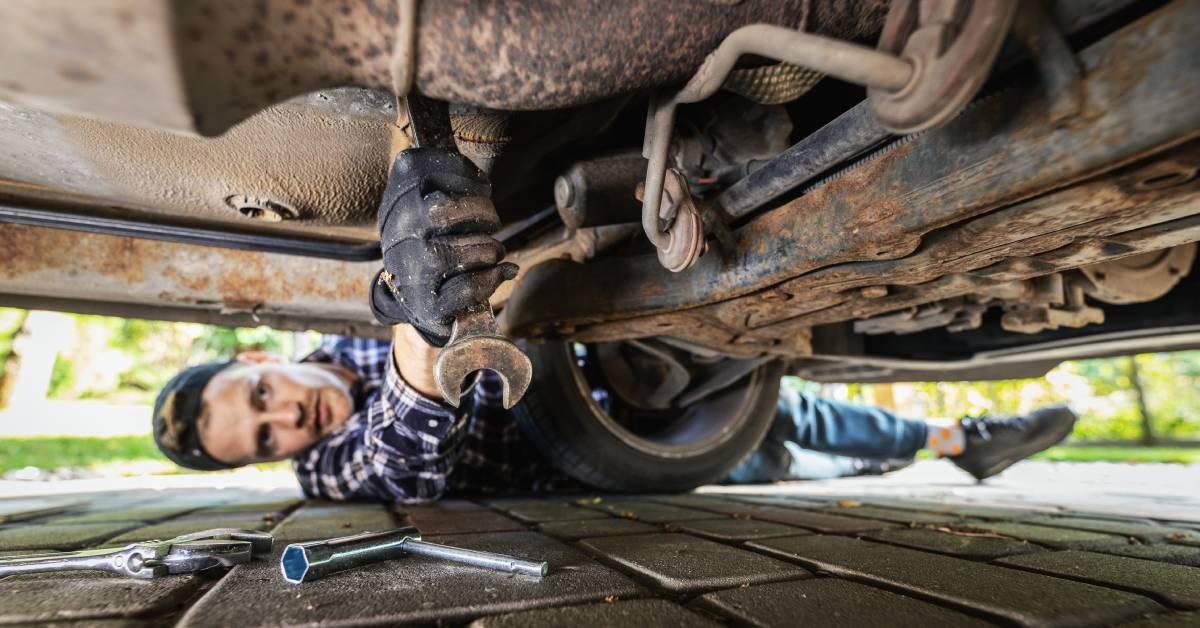 A young man laying on the ground and using a wrench to work on a vehicle's heavily rusted underside.