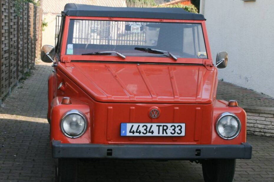 A classic orange Volkswagen Thing parked on a shaded cobblestone driveway between a brick wall and a white building.