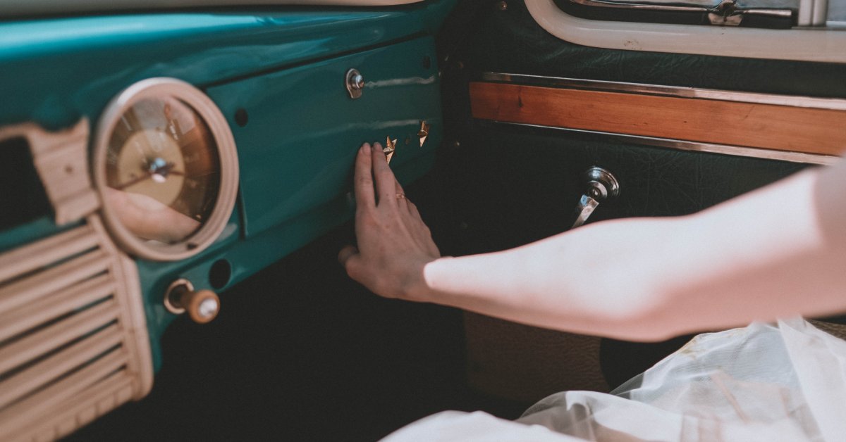 A hand touching a vintage car dashboard with a teal-green interior, cream steering wheel, and wood trim.
