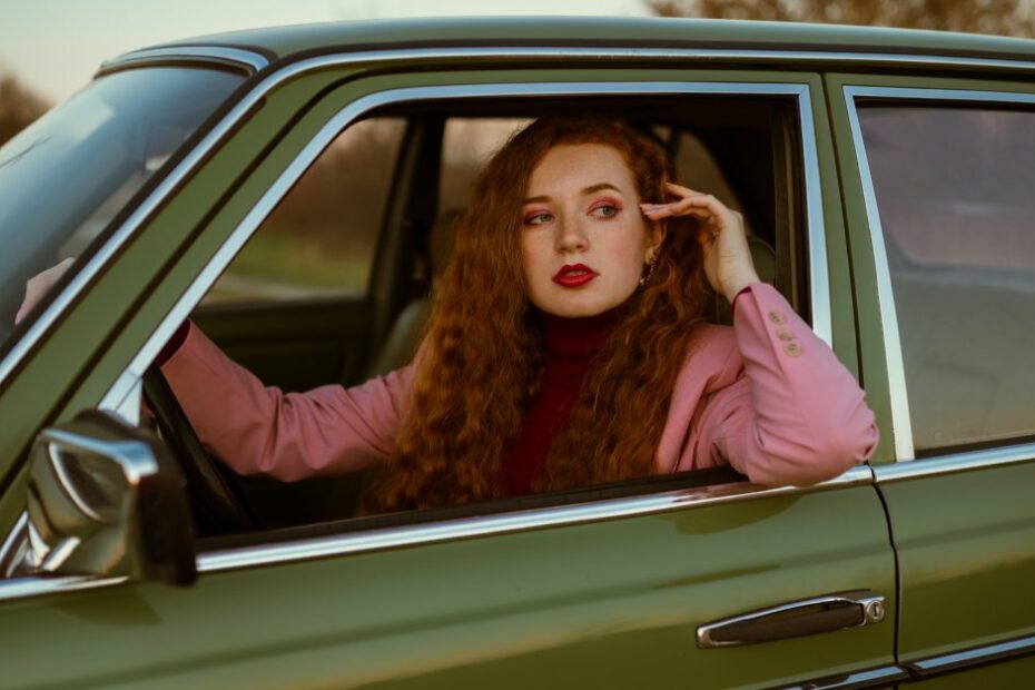 A young woman with long red hair and red lipstick sitting in the driver's seat of a vintage green car.