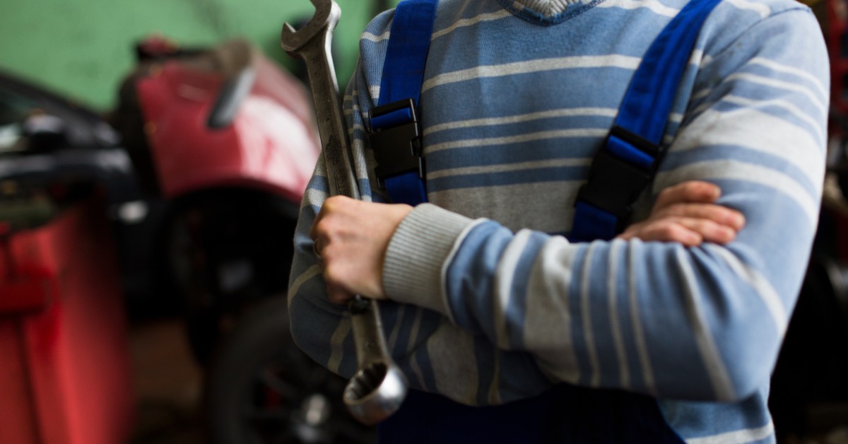 A mechanic in blue overalls stands inside an auto shop with their arms crossed and a wrench in one hand.