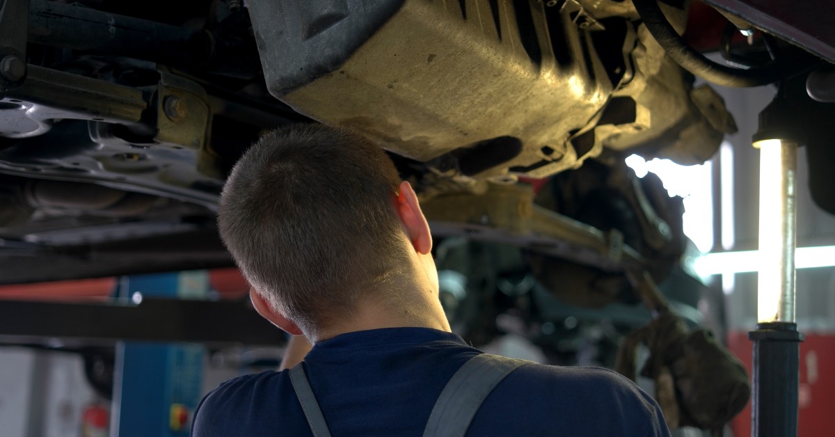 A mechanic in a blue shirt and overalls examines the underside of a lifted vehicle in an auto repair shop.
