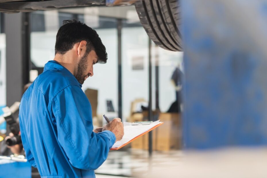 A mechanic in a blue jumpsuit stands under a lifted vehicle in an auto repair shop, writing notes on a clipboard.