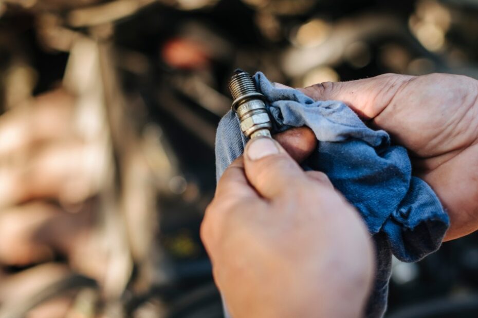 A close-up of a pair of hands cleaning a dirty spark plug with a blue rag above an exposed car engine.