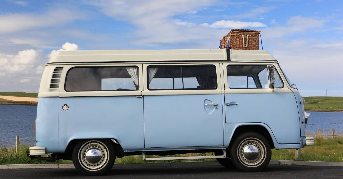 A light blue Volkswagen Bus parked on the side of the road with a wicker picnic basket strapped to its roof.