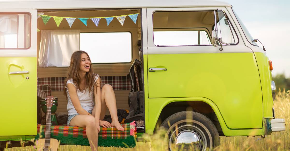 A smiling young woman with long hair sits barefoot in the open doorway of a vintage green Volkswagen Bus.