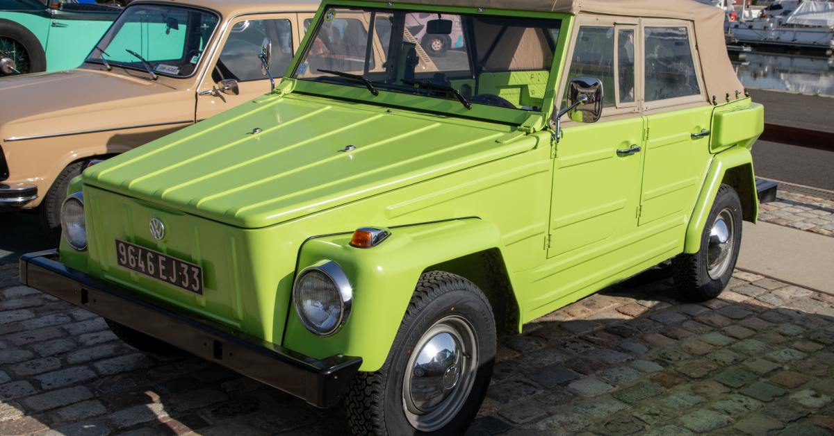 A bright green vintage Volkswagen Thing with a beige roof is parked on cobblestones at a waterfront.
