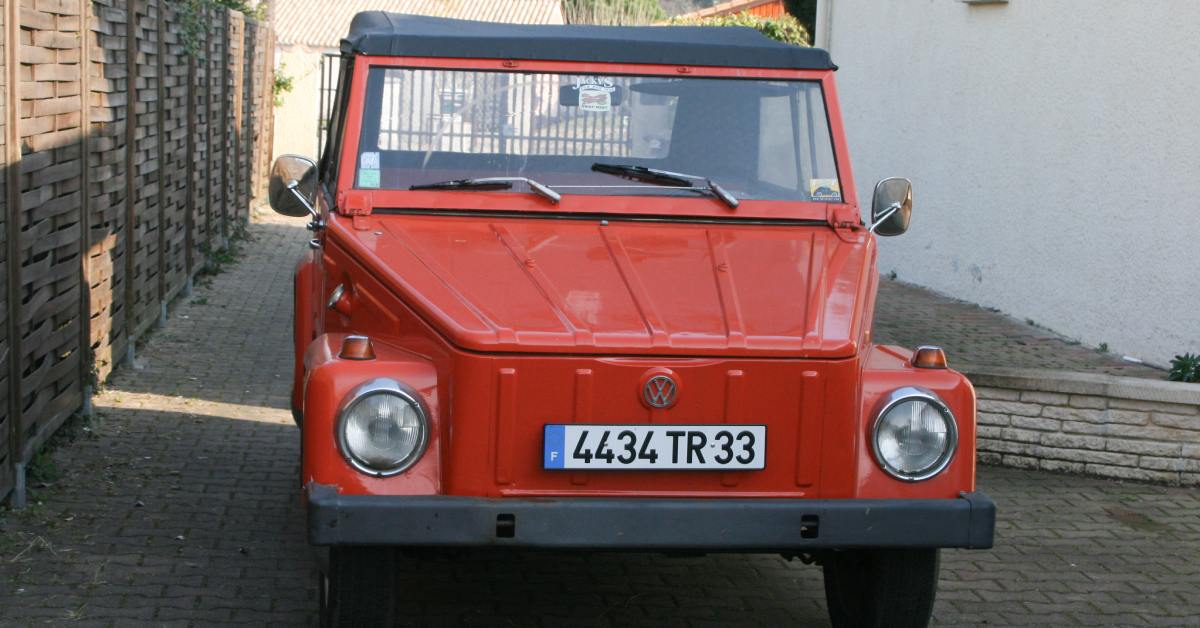 A classic orange Volkswagen Thing parked in a shaded driveway between a wooden fence and a white building.