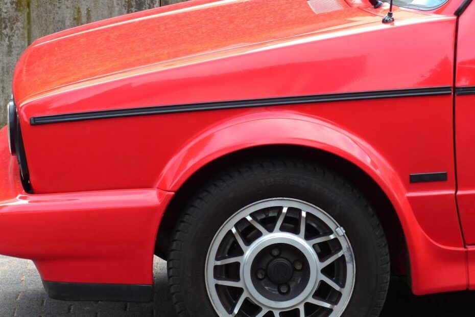 A red vintage car with spoke alloy wheels parked on a cobblestone road next to a weathered concrete wall.